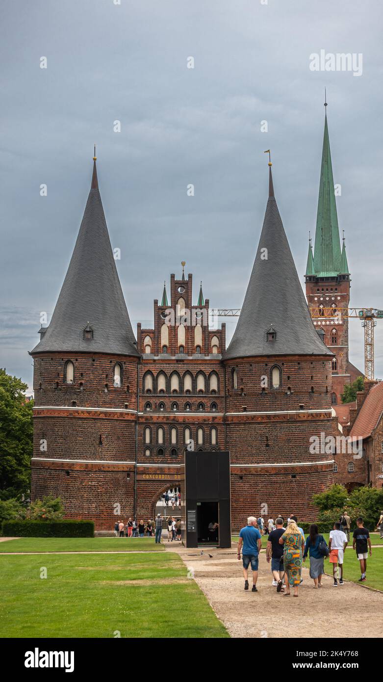 Germania, Lubecca - 13 luglio 2022: Ritratto, storica porta Holsten, Tor, con San Petri, Pietro, chiesa sul retro sotto il cielo grigio pesante. Parco verde di fronte Foto Stock