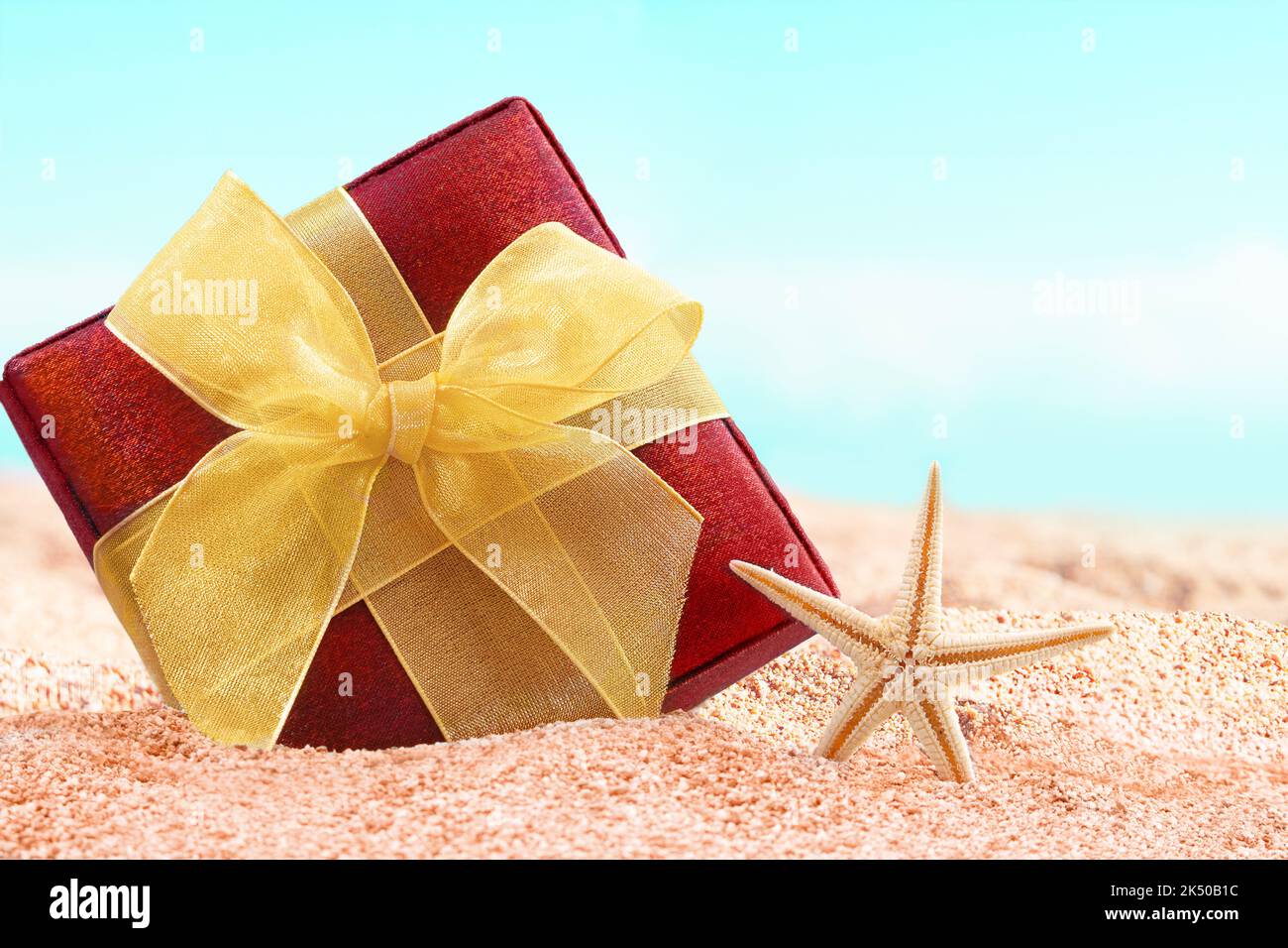 Scatola regalo rossa con arco giallo sulla spiaggia di sabbia con stelle marine. Vendita. Natale, Capodanno. Vacanze in paesi caldi Foto Stock