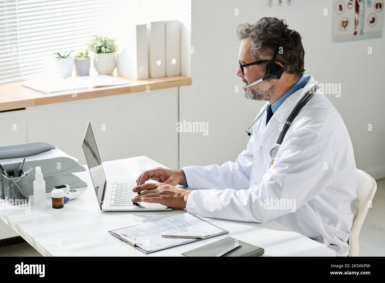 Medico maturo in cuffie a scrivere sul portatile e parlare con il paziente online sul suo posto di lavoro in ufficio Foto Stock