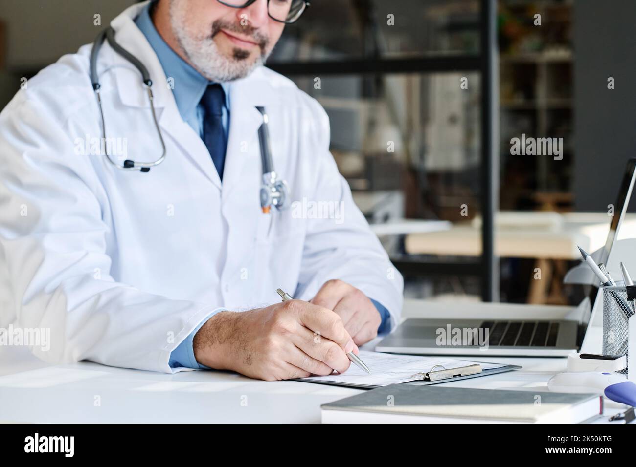 Primo piano di un medico maturo in cappotto bianco seduto a tavola in ufficio e riempiendo la scheda medica del paziente Foto Stock