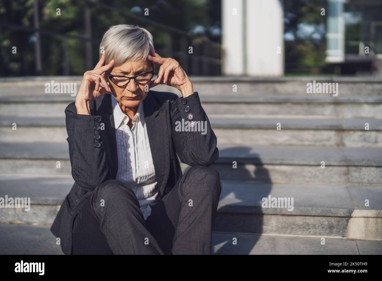 Donna d'affari anziana seduta davanti all'edificio dell'azienda. È stanca e ha mal di testa. Foto Stock
