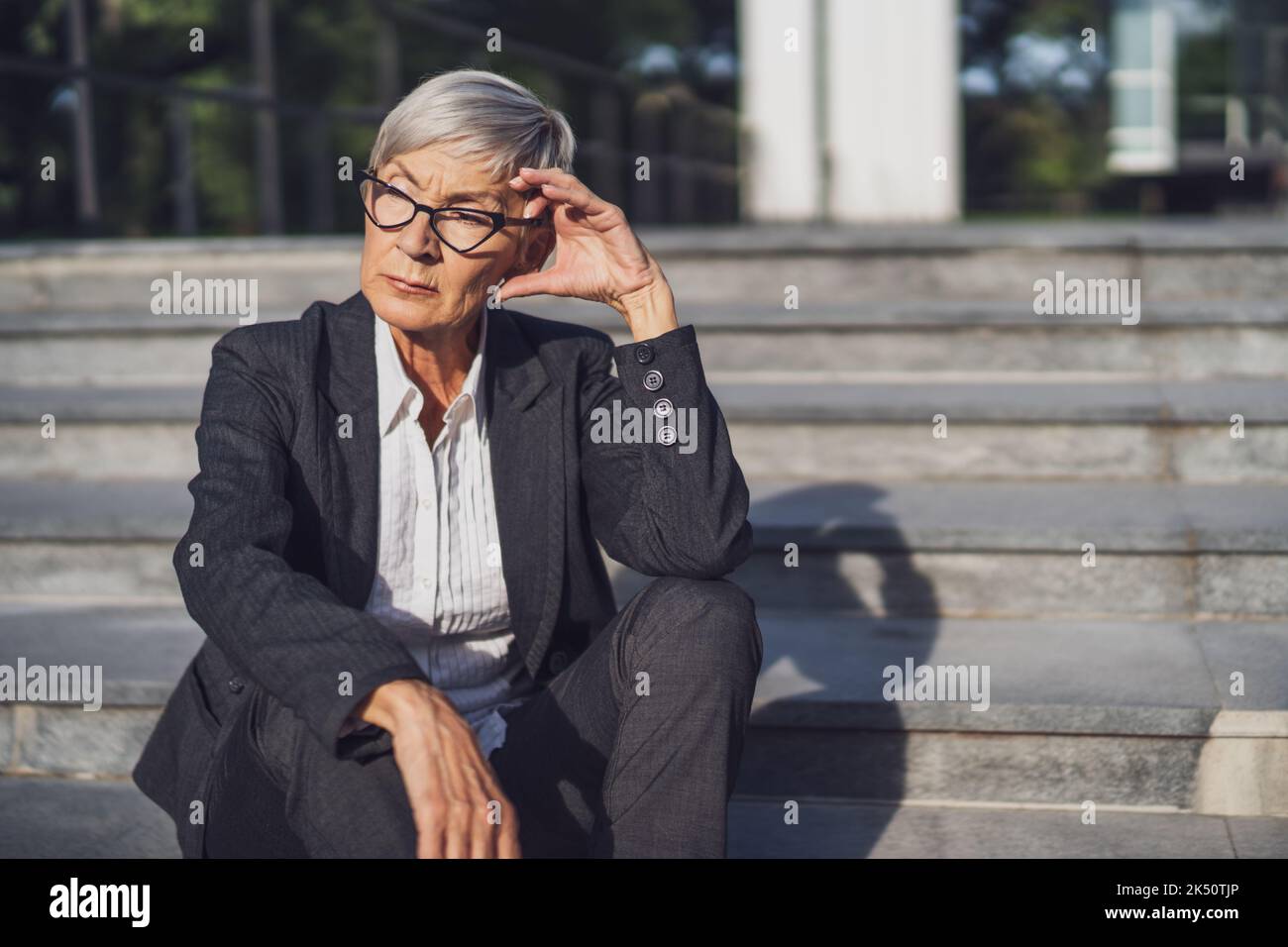Donna d'affari anziana seduta davanti all'edificio dell'azienda. È pensierosa. Foto Stock