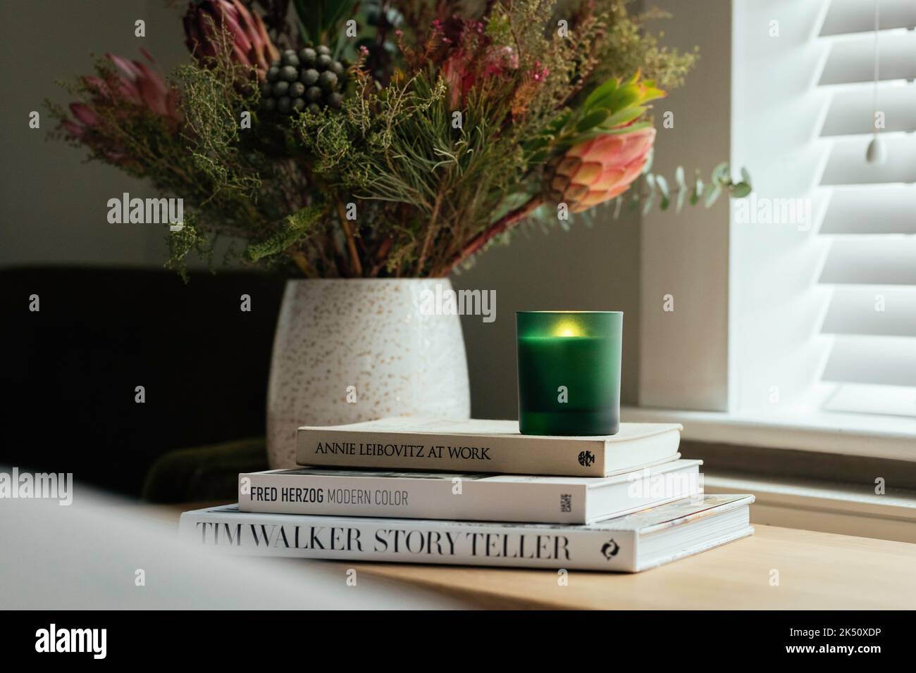 Soy Candle in un vaso di vetro verde seduto su una pila di libri in camera da letto tra fiori autunnali Foto Stock