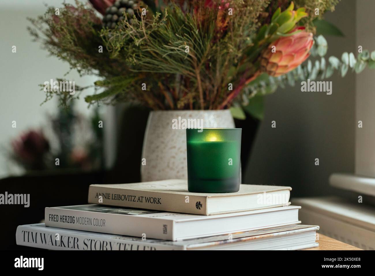 Soy Candle in un vaso di vetro verde seduto su una pila di libri in camera da letto tra fiori autunnali Foto Stock
