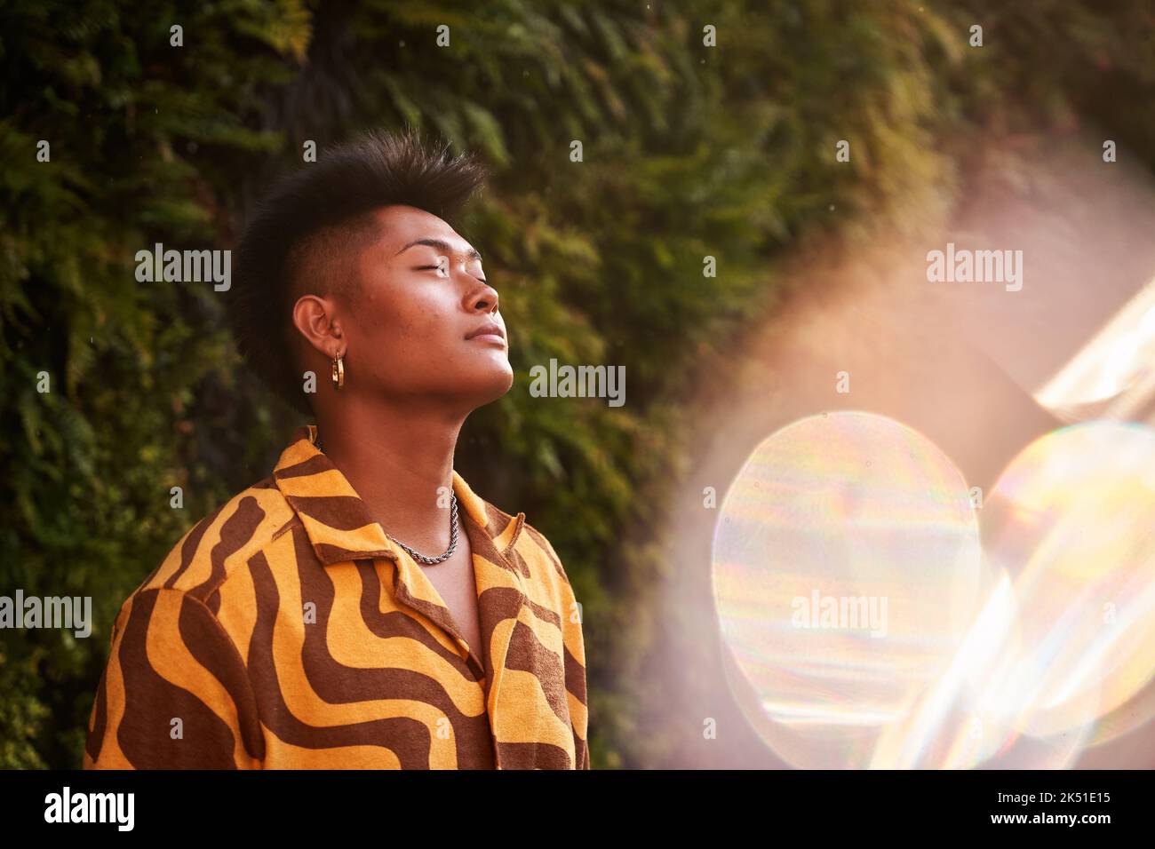 Giovane filippino da sogno non binary uomo che indossa una camicia fredda con orecchini in piedi con gli occhi chiusi nel giardino verde Foto Stock