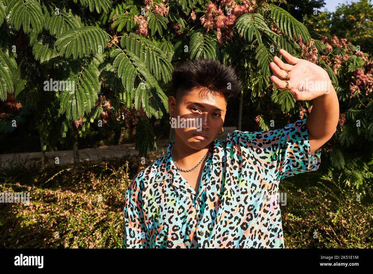 Donna asiatica transgender in elegante camicia colorata con stampa animale che protegge il viso dal sole e guarda la macchina fotografica mentre si trova vicino all'albero tropicale o Foto Stock