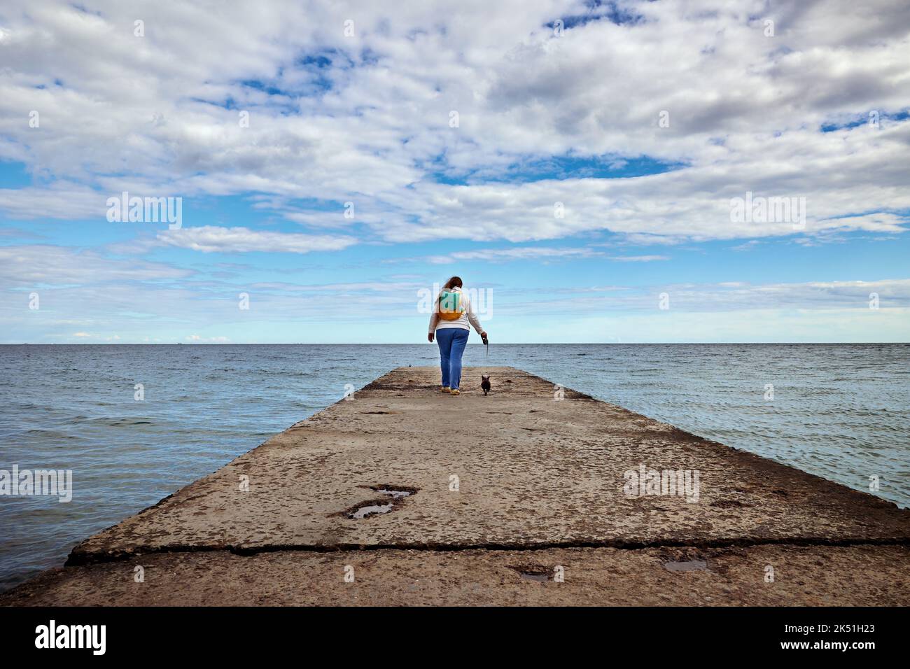 Una donna con un cane piccolo su una passeggiata al guinzaglio lungo il molo verso il cielo blu con nuvole di cirrus bianco Foto Stock