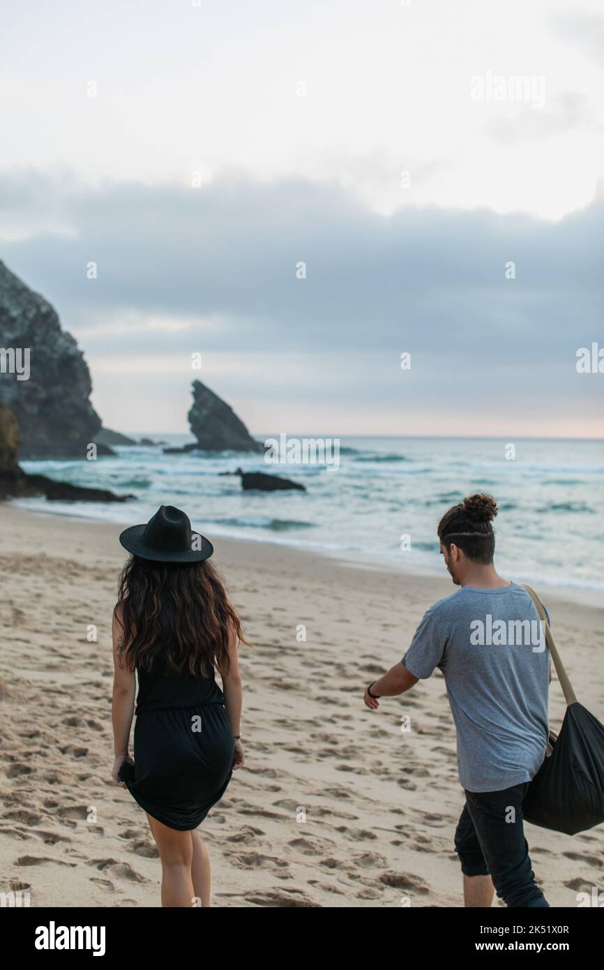 vista posteriore della donna tatuata e dell'uomo bearded con il sacchetto che cammina insieme sulla spiaggia sabbiosa, immagine di scorta Foto Stock
