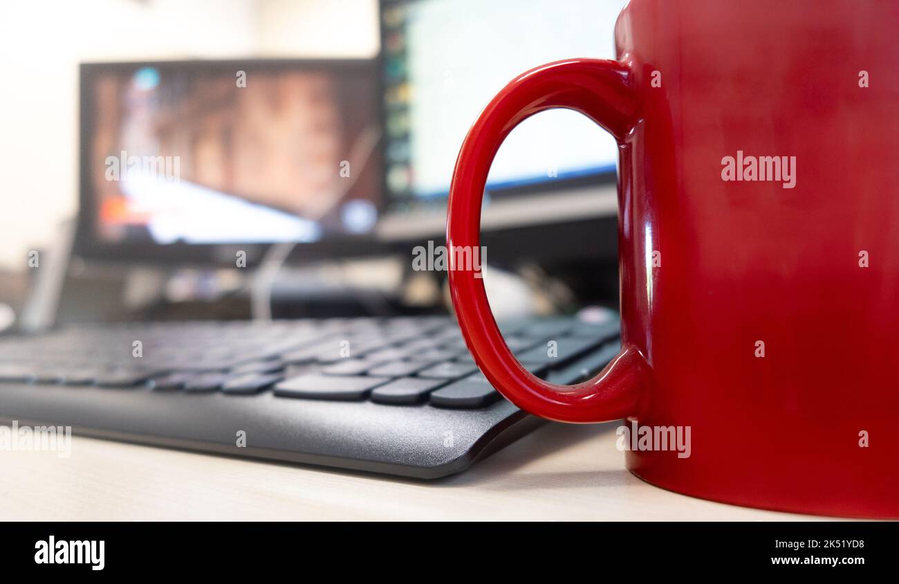Spazio di lavoro scrivania ufficio mattina vista a basso angolo primo piano della tazza di caffè rosso sfocato schermo del computer in background sul tavolo Foto Stock