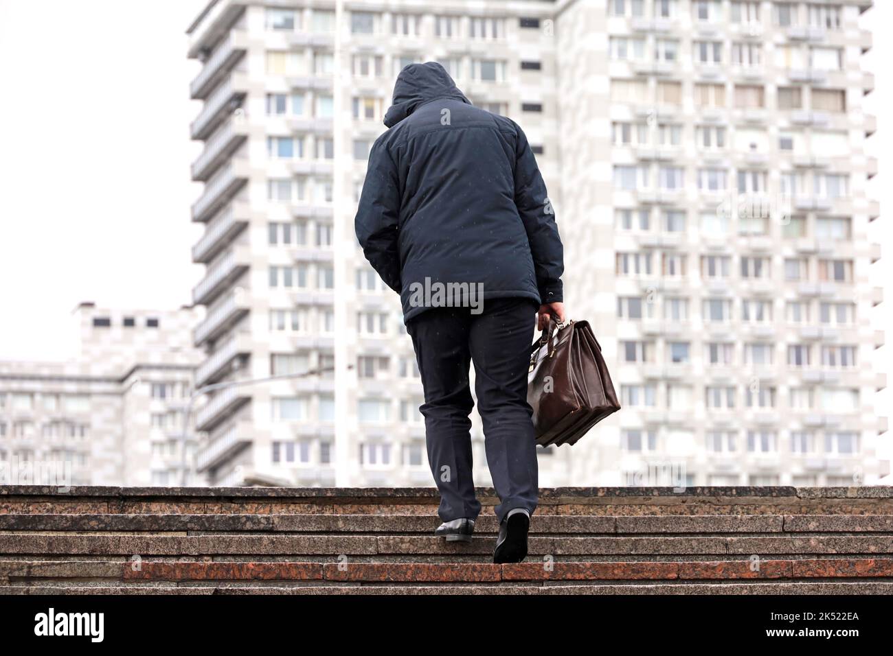 Uomo in giacca con cappuccio con valigetta in pelle che sale le scale in pietra in città durante la pioggia. Concetto di carriera, di successo, di spostamento verso l'alto Foto Stock