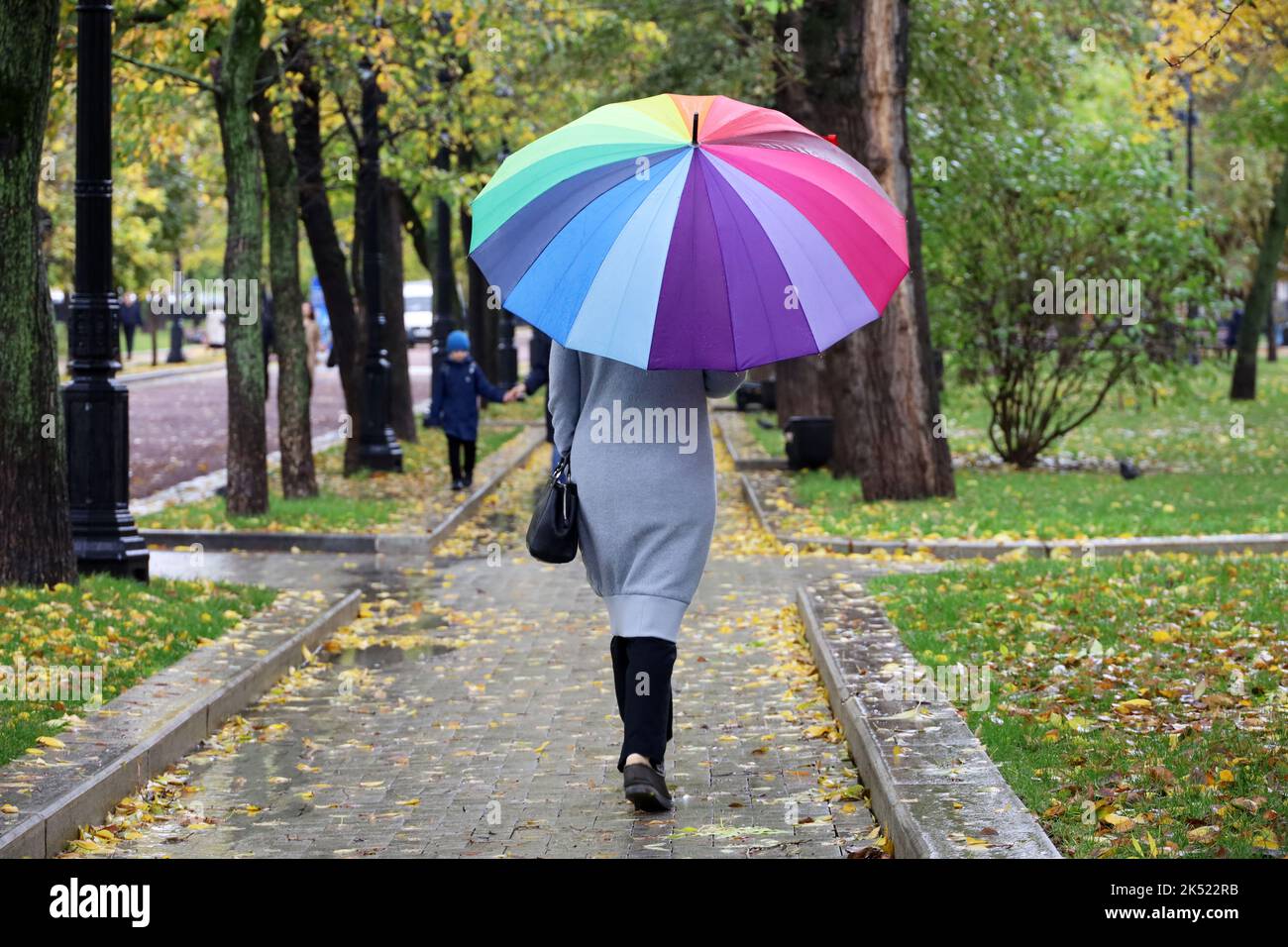 Pioggia in città, donna snella con ombrello colorato con cappotto grigio che cammina per la strada. Tempo piovoso nel parco autunnale Foto Stock