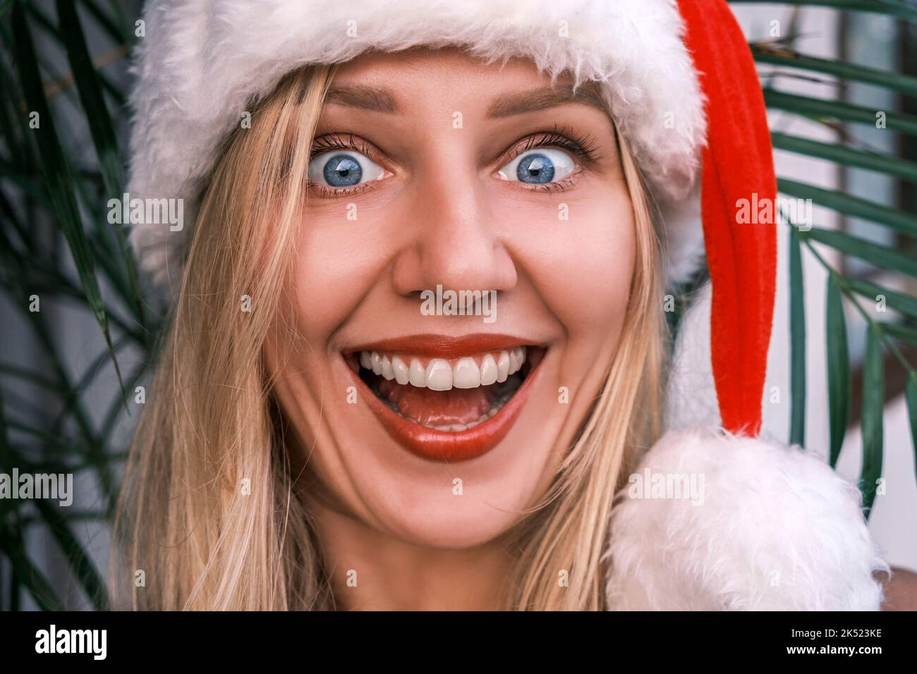 Divertente donna bionda sorridente che indossa il cappello di Santa guardando in macchina fotografica con gli occhi sorpresi. Ragazza che celebra il Natale nei tropici. Atmosfera festiva Foto Stock