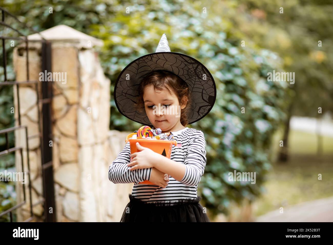 Piccola ragazza carina in costume da strega con secchio di zucca jack-o-lanterna con caramelle e dolci. Capretto trucco o trattando in festa di Halloween Foto Stock