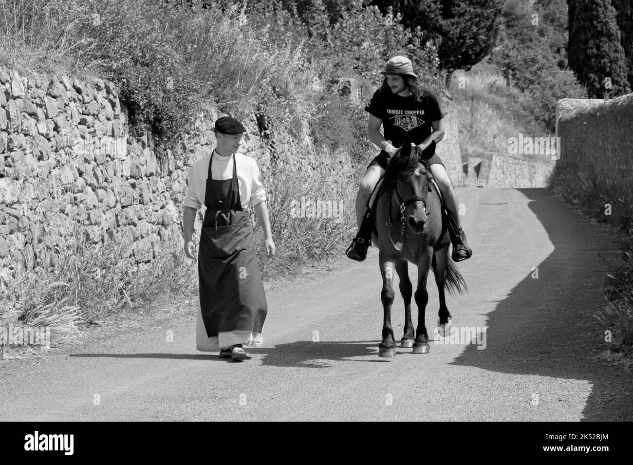 Lagrasse Aude Francia. 06.16.22 Summers mattina. Corsia di paese con muro di pietra. Giovane monaco che indossa cappello piatto e grembiule lungo sopra la sua abitudine cammina e chiacchierare Foto Stock