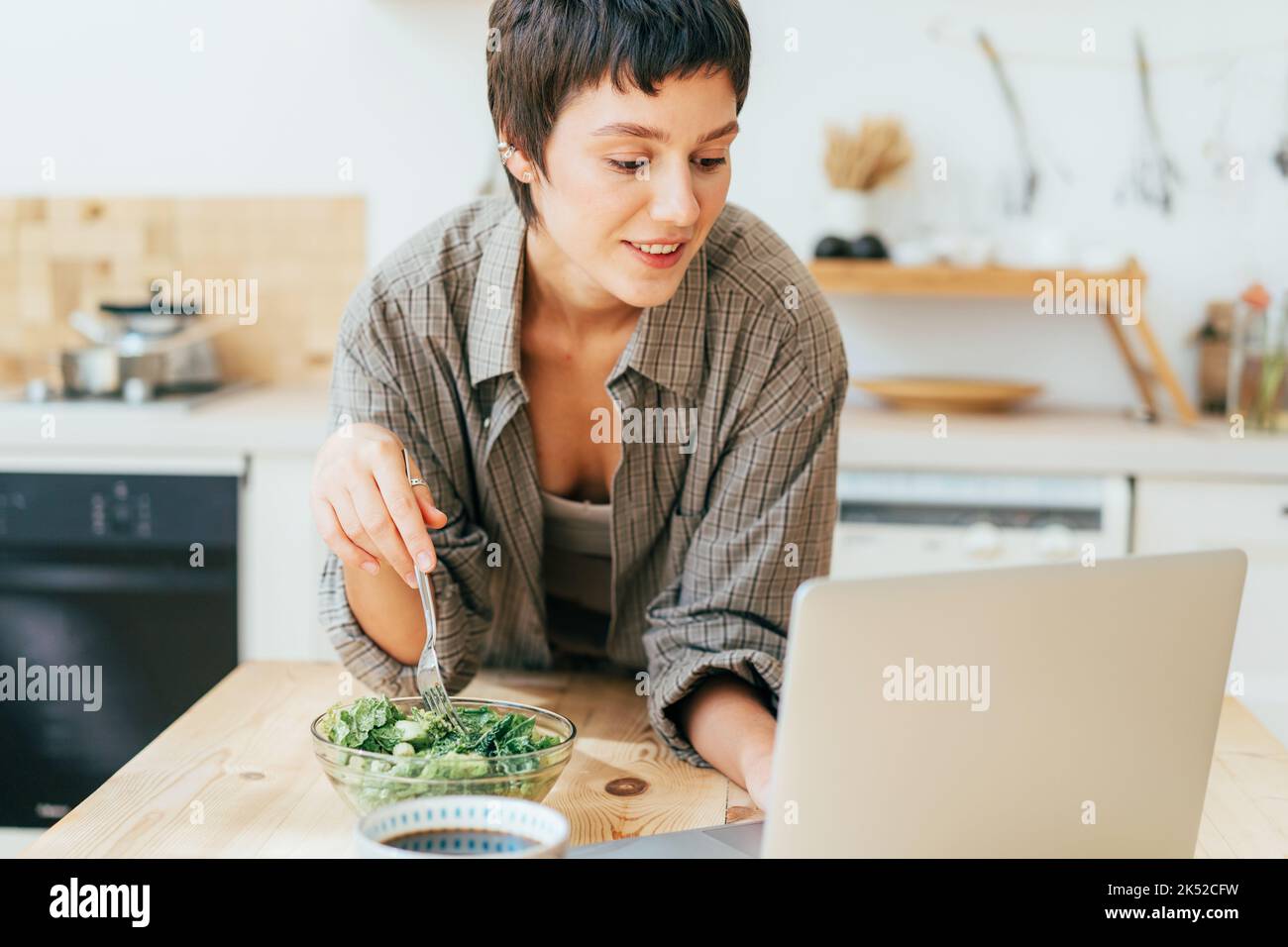 Una giovane donna attraente in cucina mangia un'insalata verde sana e lavora su un computer portatile. Foto Stock