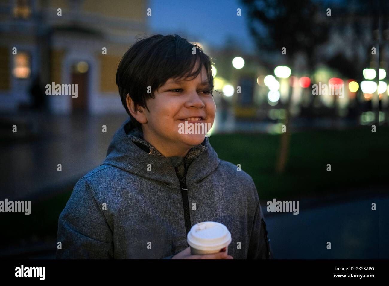 Un adolescente con emozioni positive e una tazza di carta in mano sulla strada in una serata autunno scuro sullo sfondo delle luci della città. Foto Stock