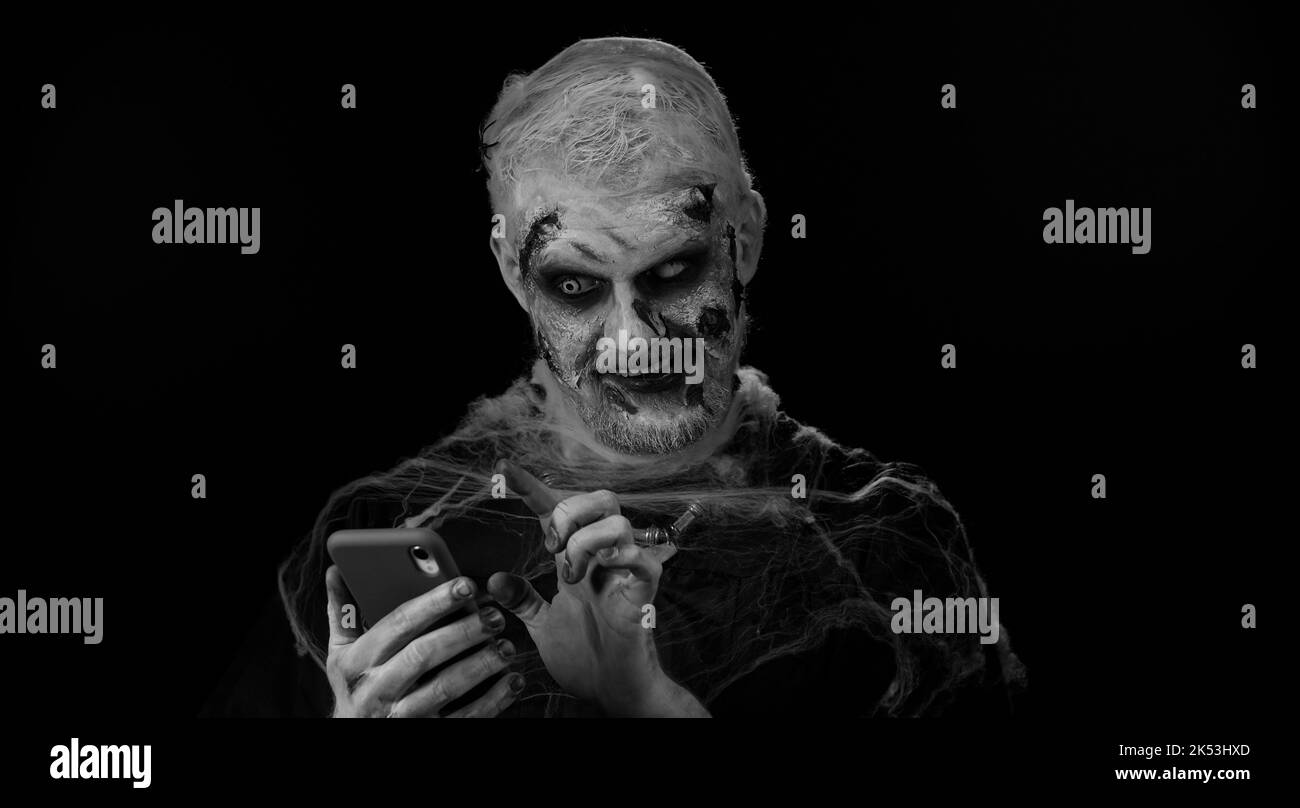 Spaventoso uomo con Halloween zombie sanguinoso ferito trucco utilizzando il telefono cellulare digitando nuovo post sul web, sms messaggio, navigazione, dipendenza di social network. Un sinistro ragazzo non morto su sfondo nero parete Foto Stock