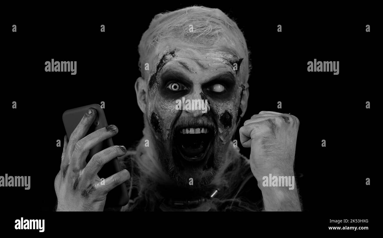Felice eccitato sinistro uomo in costume di carnevale di Halloween pazzo zombie visualizzazione smartphone sinceramente esultante vittoria, ricevendo messaggio con buone notizie, fortuna successo. Cosplay ferito non morto Foto Stock
