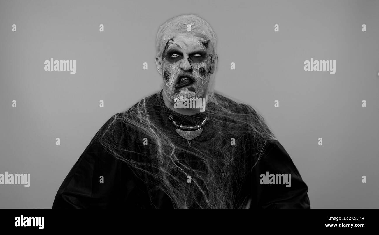 Uomo sinistro con orribile spaventoso trucco zombie Halloween rendendo divertenti espressioni facciali sciocco e grimacing, imbrogliando intorno mostrando lingua. Ragazzo morto con ferite sanguinose cicatrici faccia. Sfondo nero Foto Stock