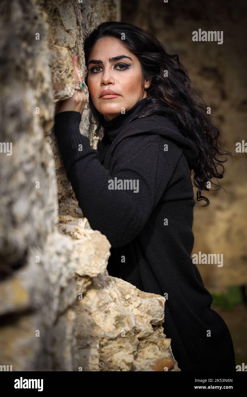 Ritratto di bruna donna persiana con capelli lunghi in abito nero in piedi contro muro rovinato con un look attento. Foto Stock