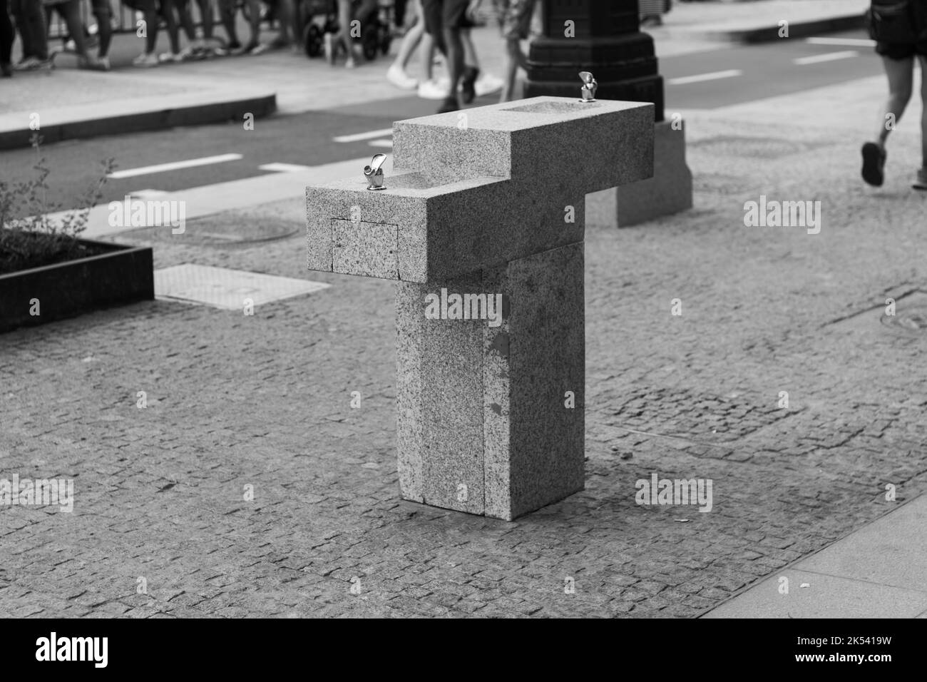Una scala di grigi di una fontana di bere geometrica e una strada asfaltata e marciapiede con persone sullo sfondo. Foto Stock