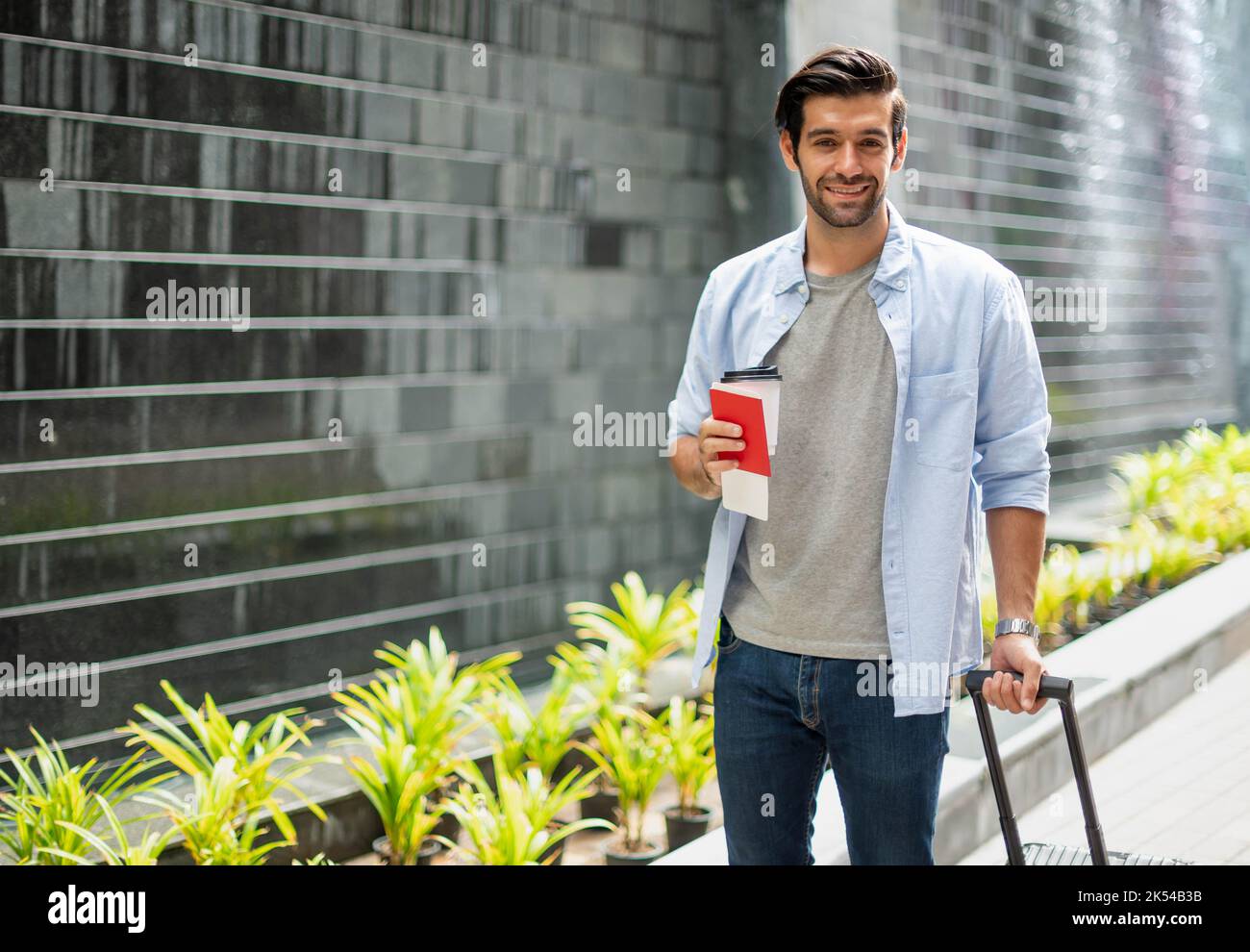 Giovane uomo caucasico che tiene la tazza da caffè e tira la valigia aspettando il suo amico per viaggiare insieme. Foto Stock