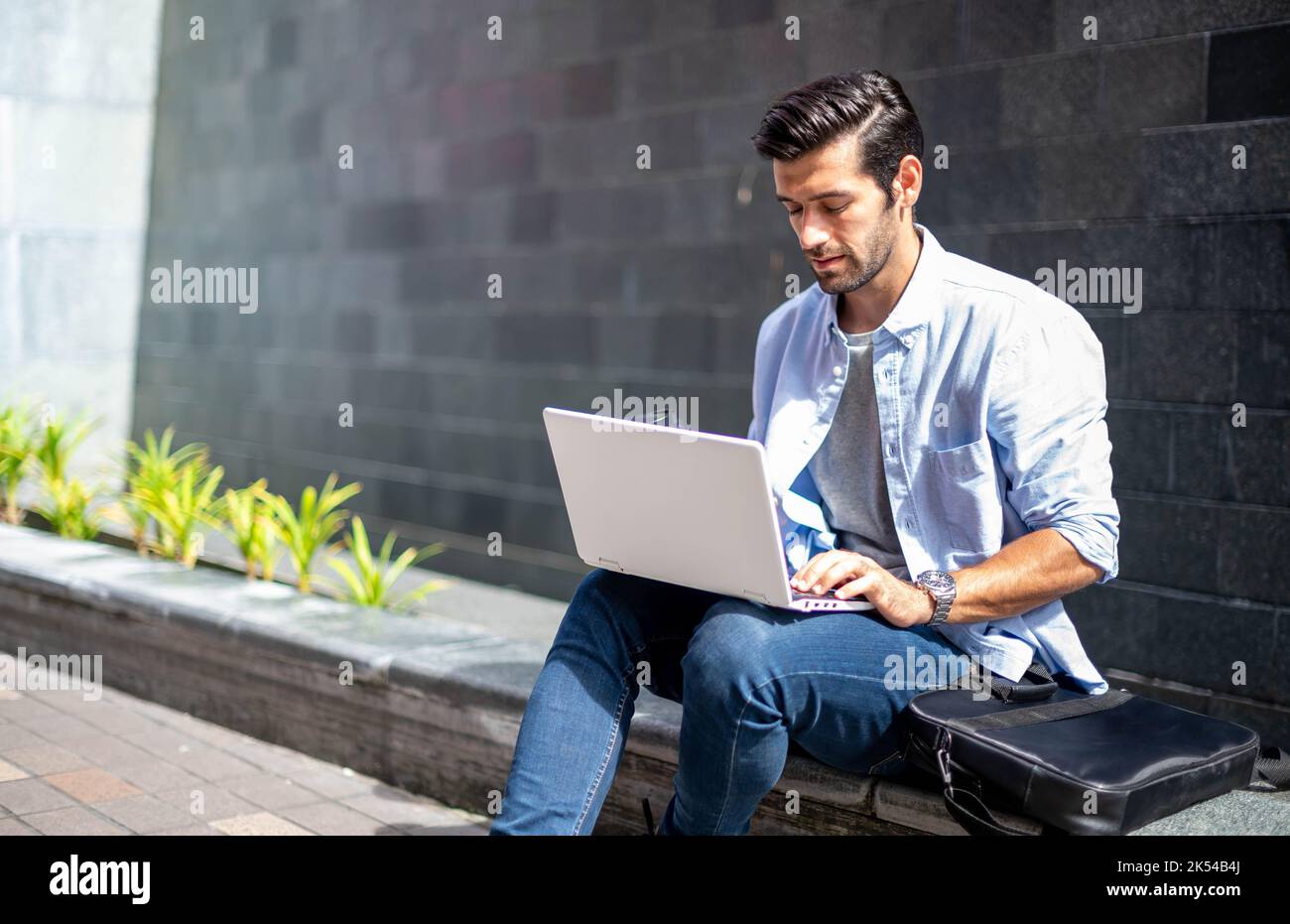 Giovane uomo caucasico seduto e lavorando a progetto freelance utilizzando computer portatile in ufficio esterno. Foto Stock