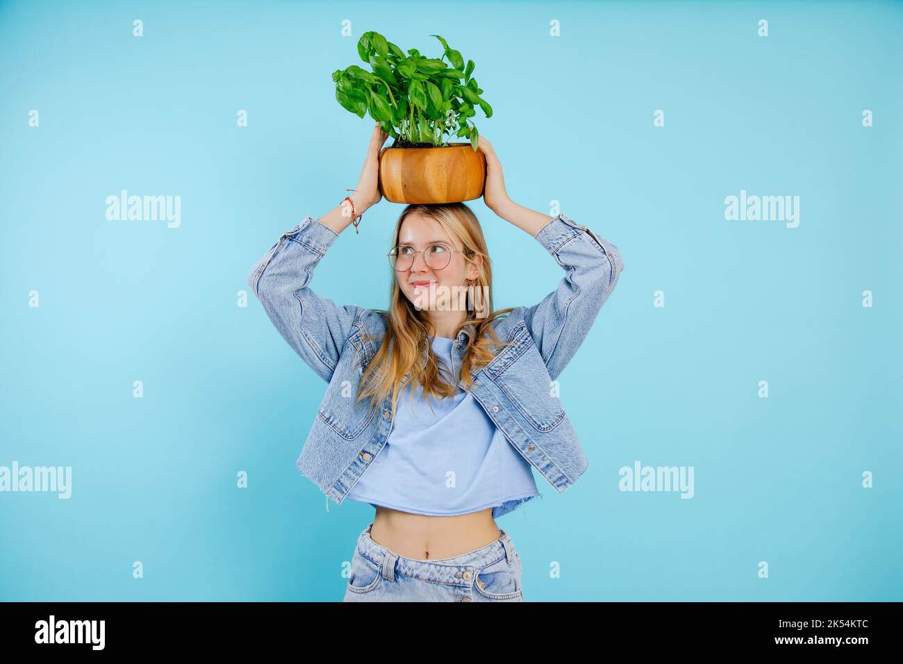 Bella ragazza adolescente tenere sulla testa pianta verde casa in pentola di legno su sfondo blu vuoto, spazio copia libero. Foto di giovane signora piuttosto sottile che cresce Foto Stock