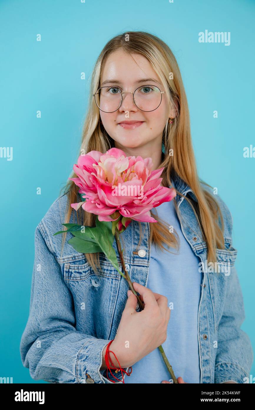 Ragazza adolescente cute che tiene grande fiore fresco rosa fragrante con gambo lungo su fondo blu vuoto. Felice giovane donna in occhiali con sorriso godere la bellezza Foto Stock