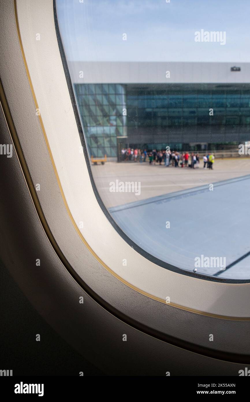Vista da una finestra di un aereo in un aeroporto, con una coda di persone in distanza lasciando le partenze per il loro aereo. Foto Stock