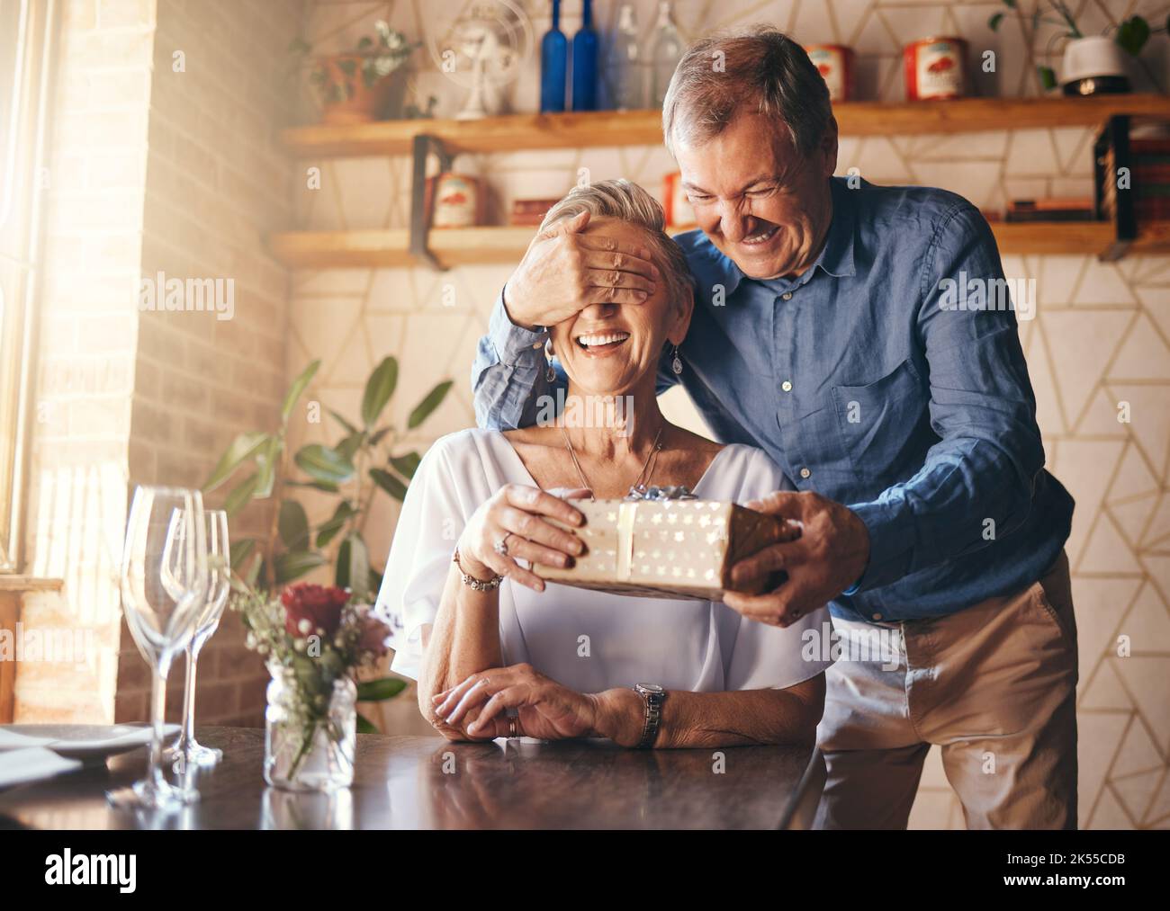 Regalo, sorpresa e coppia anziana al ristorante per la festa della pensione o buon compleanno con bicchiere di vino, regalo e rose. Donna anziana con Foto Stock