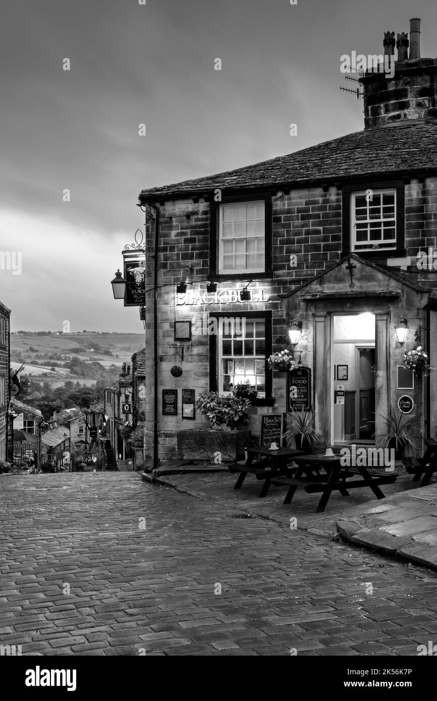 Haworth Main Street (collina ripida, vecchi edifici, storico villaggio delle sorelle di Bronte, pub di grado 2, cielo nuvoloso grigio scuro) - West Yorkshire, Inghilterra, Regno Unito. Foto Stock