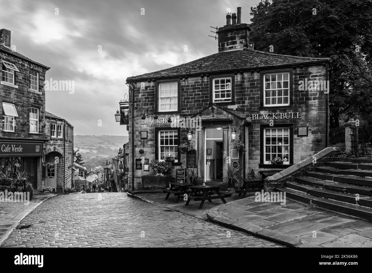 Haworth Main Street (collina ripida, vecchi edifici, storico villaggio delle sorelle di Bronte, pub di grado 2, cielo nuvoloso grigio scuro) - West Yorkshire, Inghilterra, Regno Unito. Foto Stock
