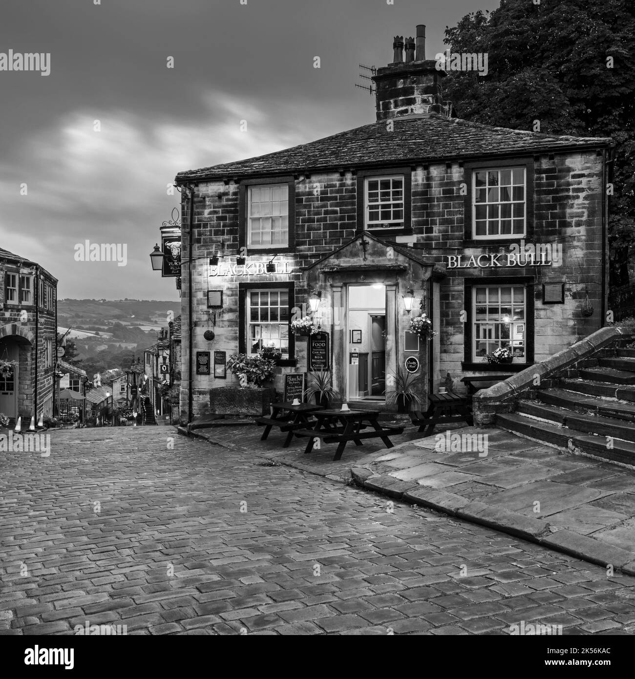 Haworth Main Street (collina ripida, vecchi edifici, storico villaggio delle sorelle di Bronte, pub di grado 2, cielo nuvoloso grigio scuro) - West Yorkshire, Inghilterra, Regno Unito. Foto Stock
