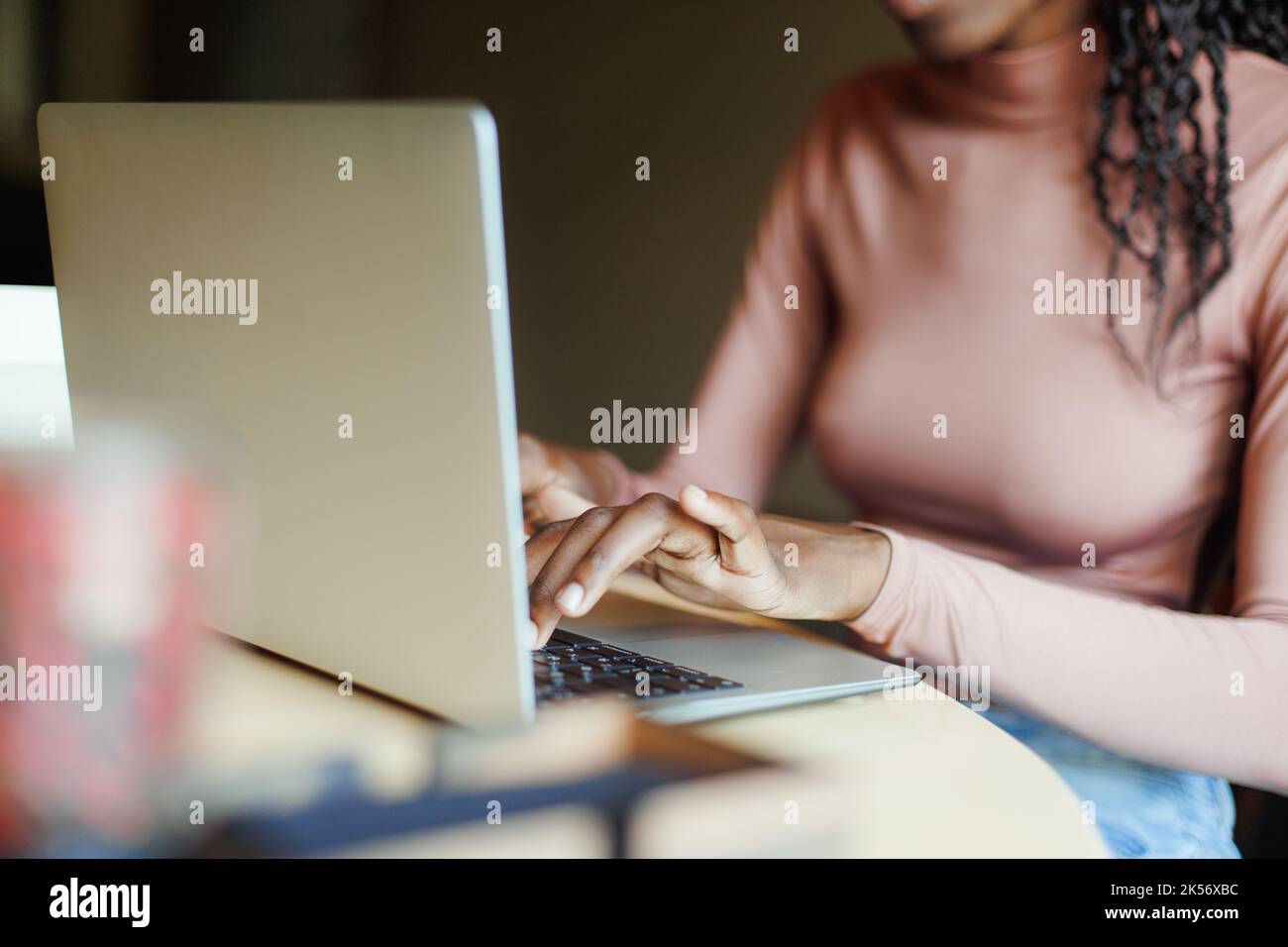 Foto ritagliata di una donna afro-americana seduta a un tavolo di legno beige, lavorando a scrivere utilizzando su un computer portatile grigio. Tecnologia. Foto Stock