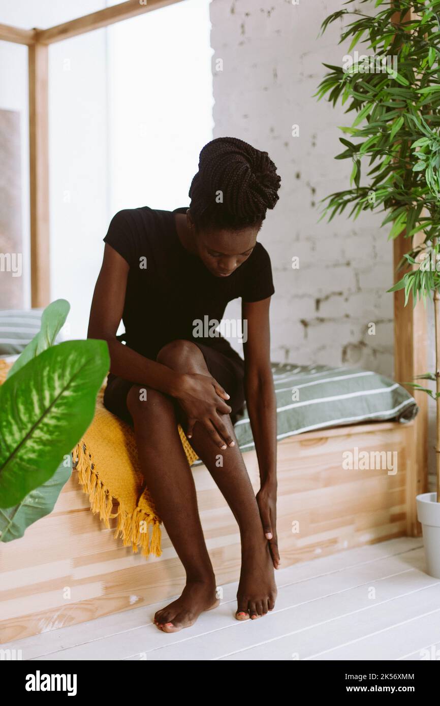 Bella donna afroamericana con dreadlocks seduta sul letto in studio decorato e massaggiante piede a mano. Persona nera che ha lesioni sportive, muscolo Foto Stock