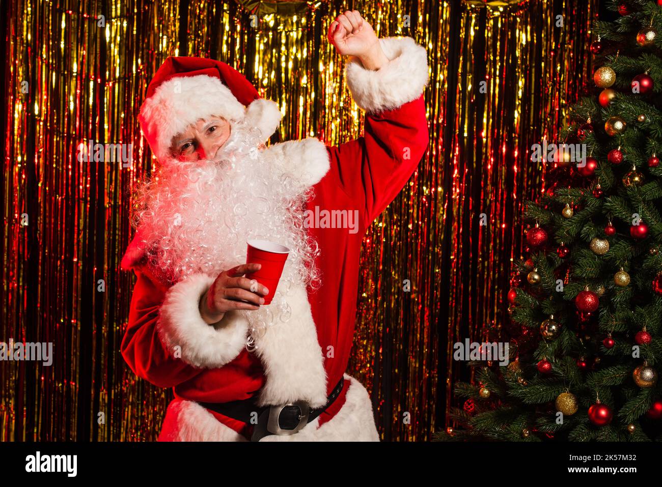 Eccitato babbo natale che tiene la tazza di plastica vicino all'albero di natale e tinsel, immagine di scorta Foto Stock