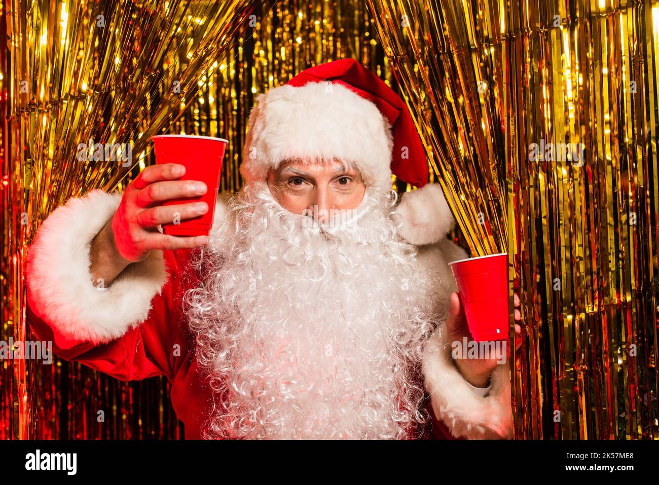 babbo natale in costume che tiene le tazze di plastica e che guarda la macchina fotografica vicino al tinsel, immagine di scorta Foto Stock