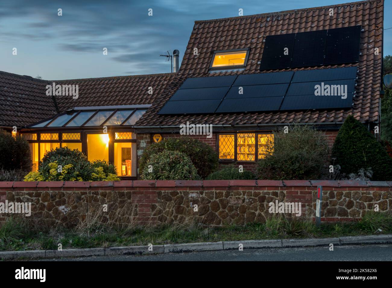 Luci che bruciano al crepuscolo in un cottage di campagna Norfolk con celle PV sul tetto. Utilizzo di energia solare risparmiata dall'immagazzinamento della batteria. Foto Stock