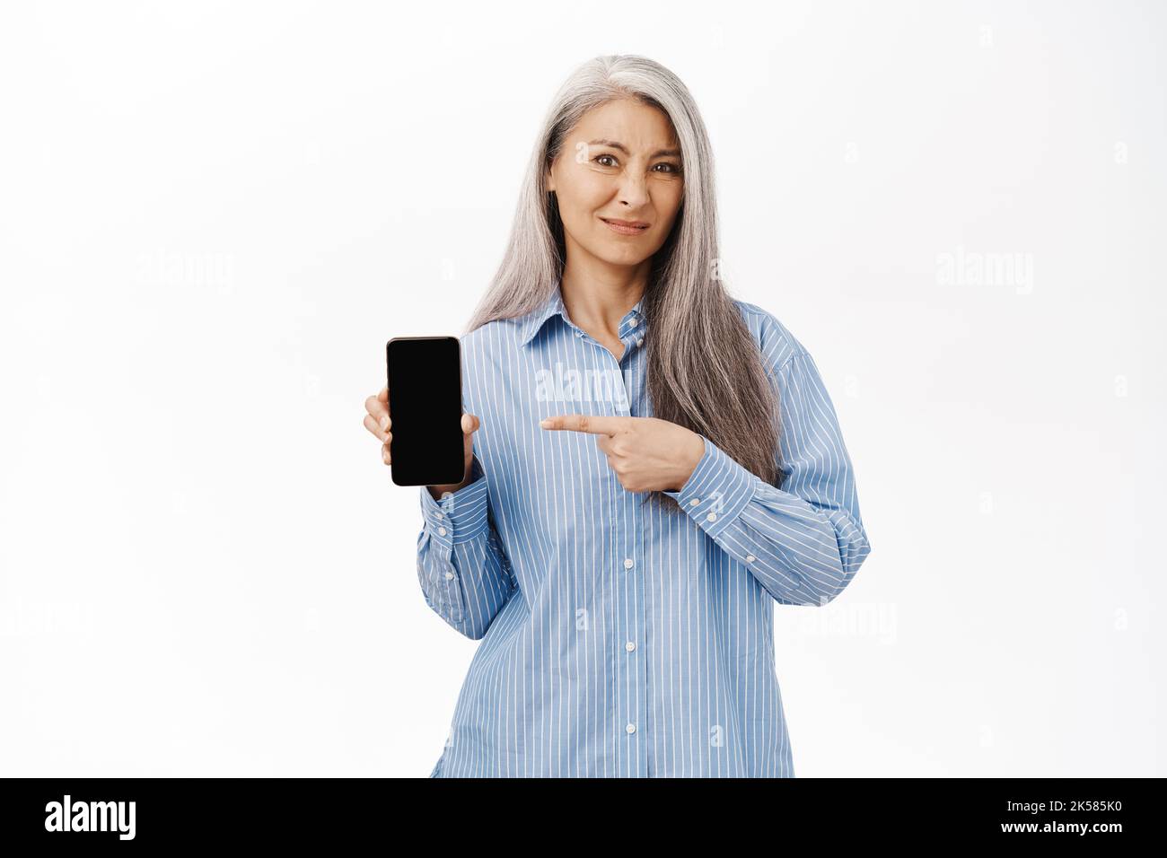 Donna asiatica anziana che punta il dito sullo schermo del telefono cellulare con il viso deluso, l'applicazione non piace o negozio web, in piedi su sfondo bianco Foto Stock