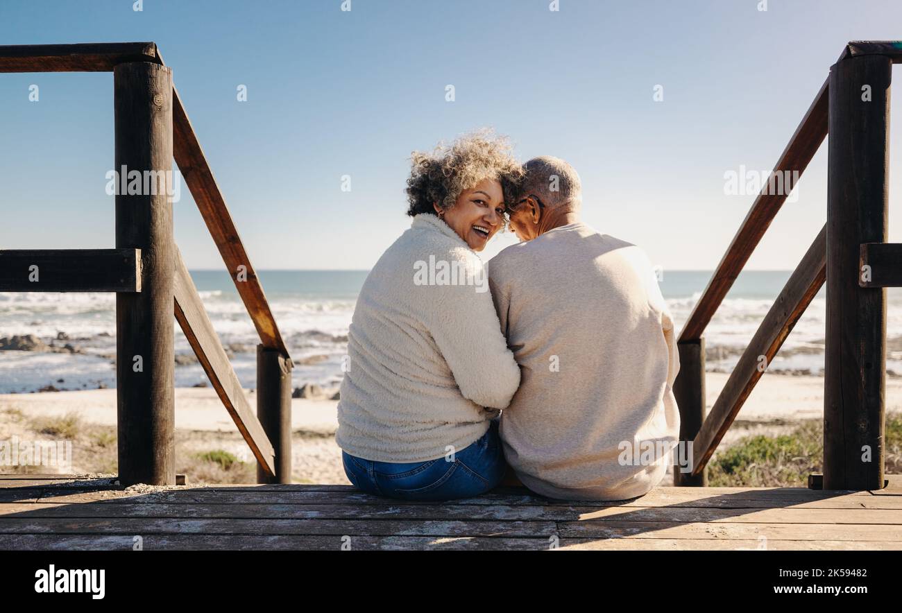 Donna anziana allegra sorridente alla macchina fotografica mentre si siede su un ponte di legno sul mare con suo marito. Coppia anziana in pensione che spende un certo ti di qualità Foto Stock