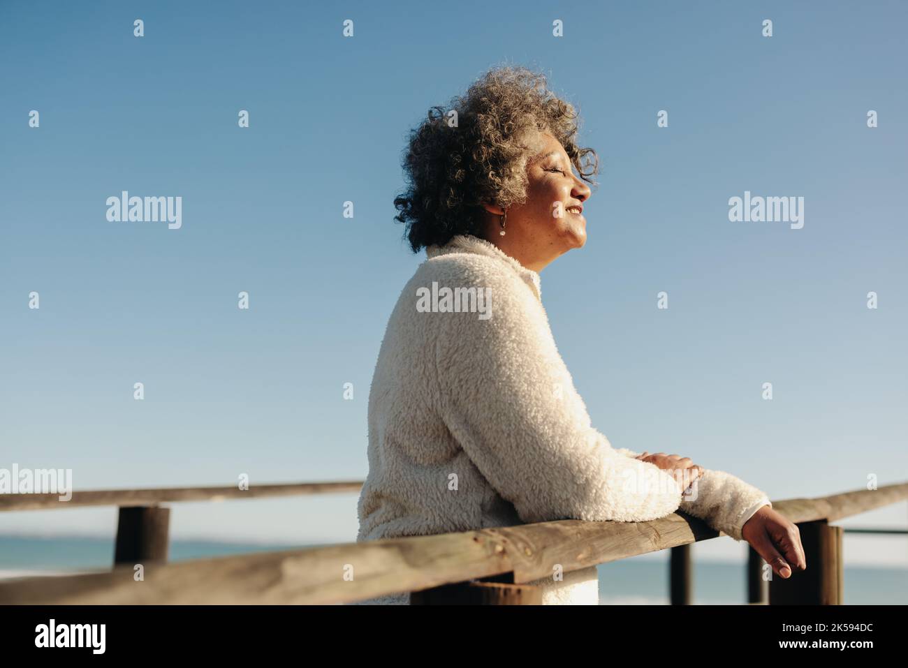 Godersi il sole in spiaggia. Donna anziana felice che ottiene un po 'di luce del sole mentre si trova su una passerella vicino all'oceano. Donna anziana allegra che prende Foto Stock