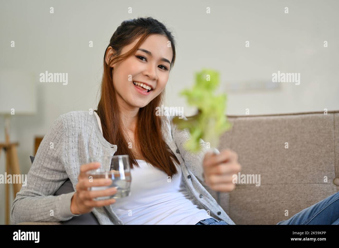 Giovane donna asiatica dall'aspetto bello seduta sul divano in soggiorno, con un bicchiere d'acqua e una foglia di lattuga verde. Concetto di stile di vita sano Foto Stock