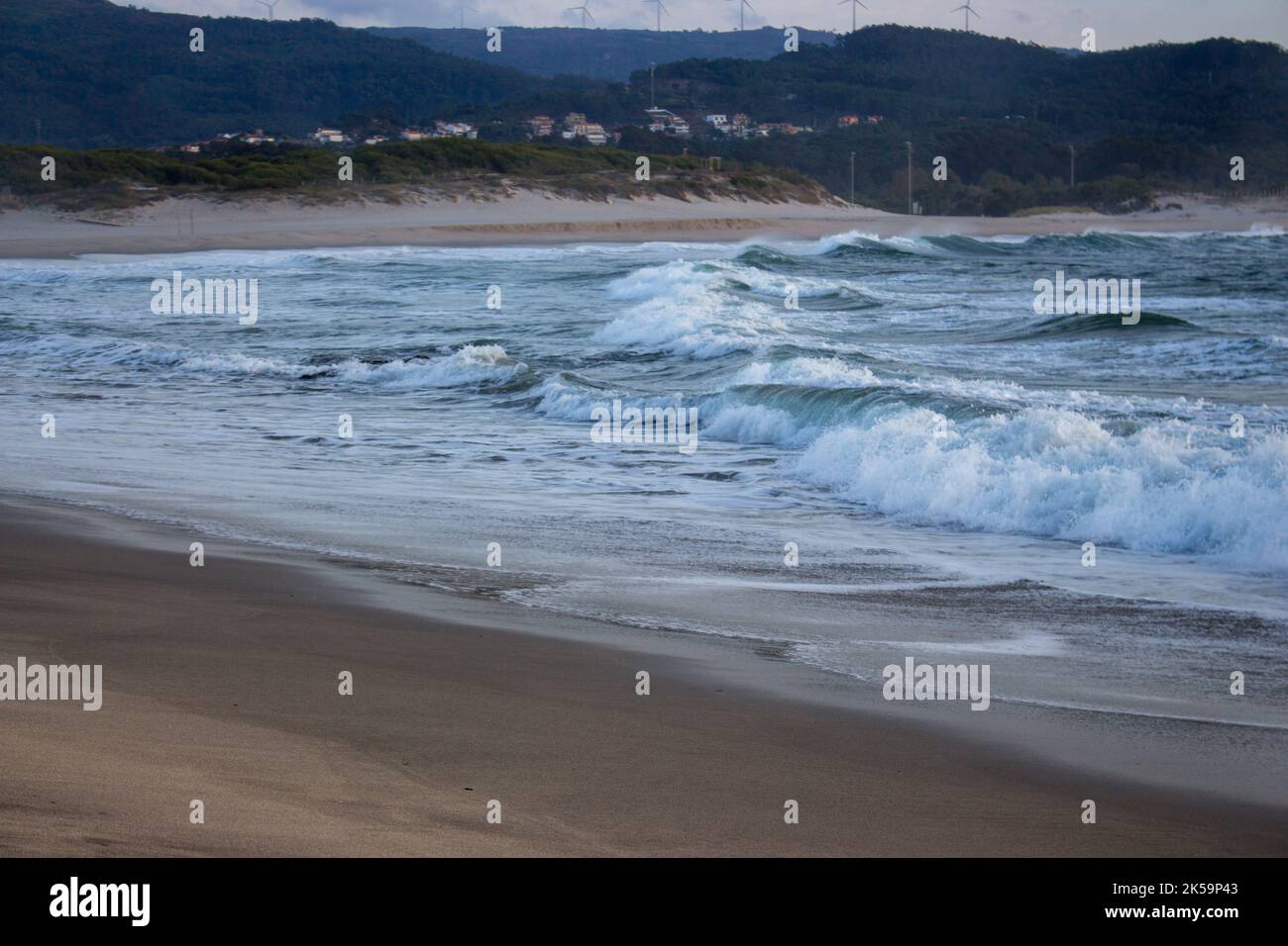 Onde dell'oceano schiumose che si infrangono sulla spiaggia sabbiosa al crepuscolo serale. Surf sulla spiaggia con villaggio e colline sullo sfondo. Bassa stagione di surf. Foto Stock
