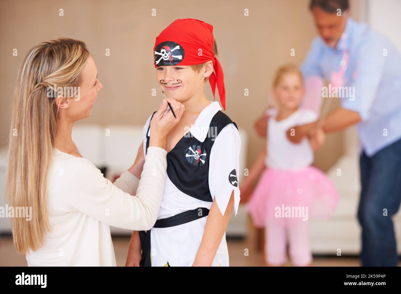 Mamma e le sue meravigliose idee. Un ragazzo sorridente in piedi di fronte alla madre che sta dipingendo un baffo sul suo viso con padre e sorella nel Foto Stock