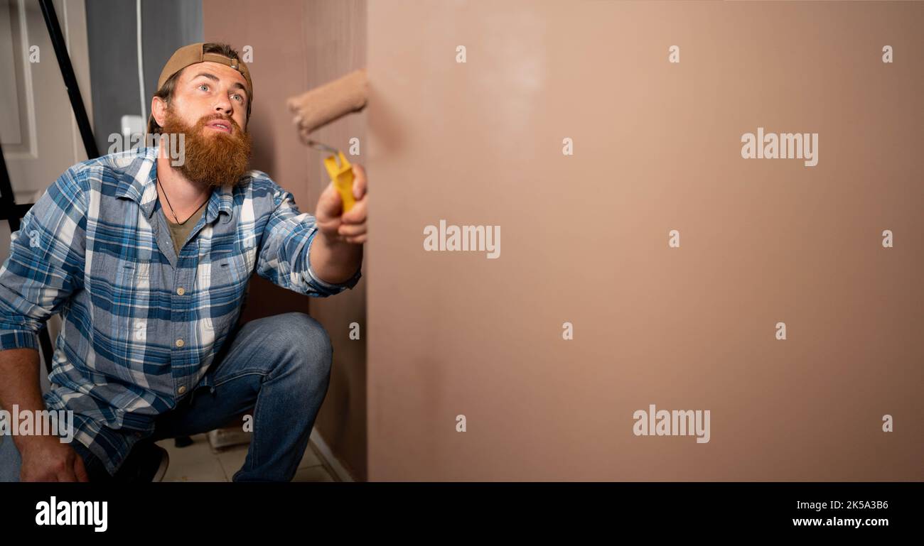 Il pittore bearded dipinge le pareti con rullo di vernice mentre fa la ristrutturazione della casa Foto Stock