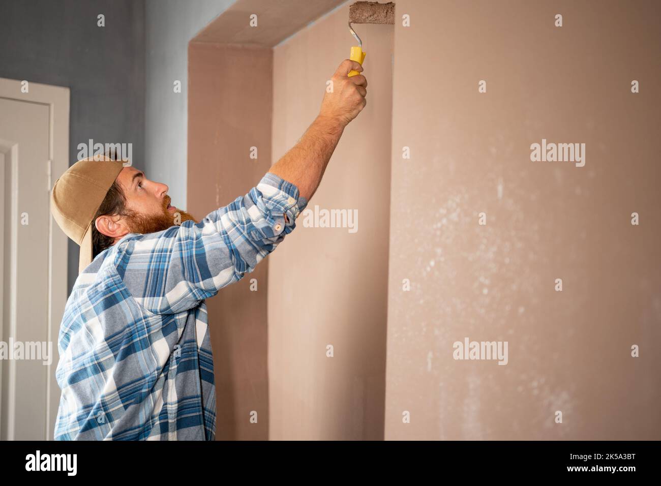 Pittore bearded dipingere un muro con un rullo di pittura su un ampio spazio vuoto. Uomo durante i lavori di ristrutturazione Foto Stock