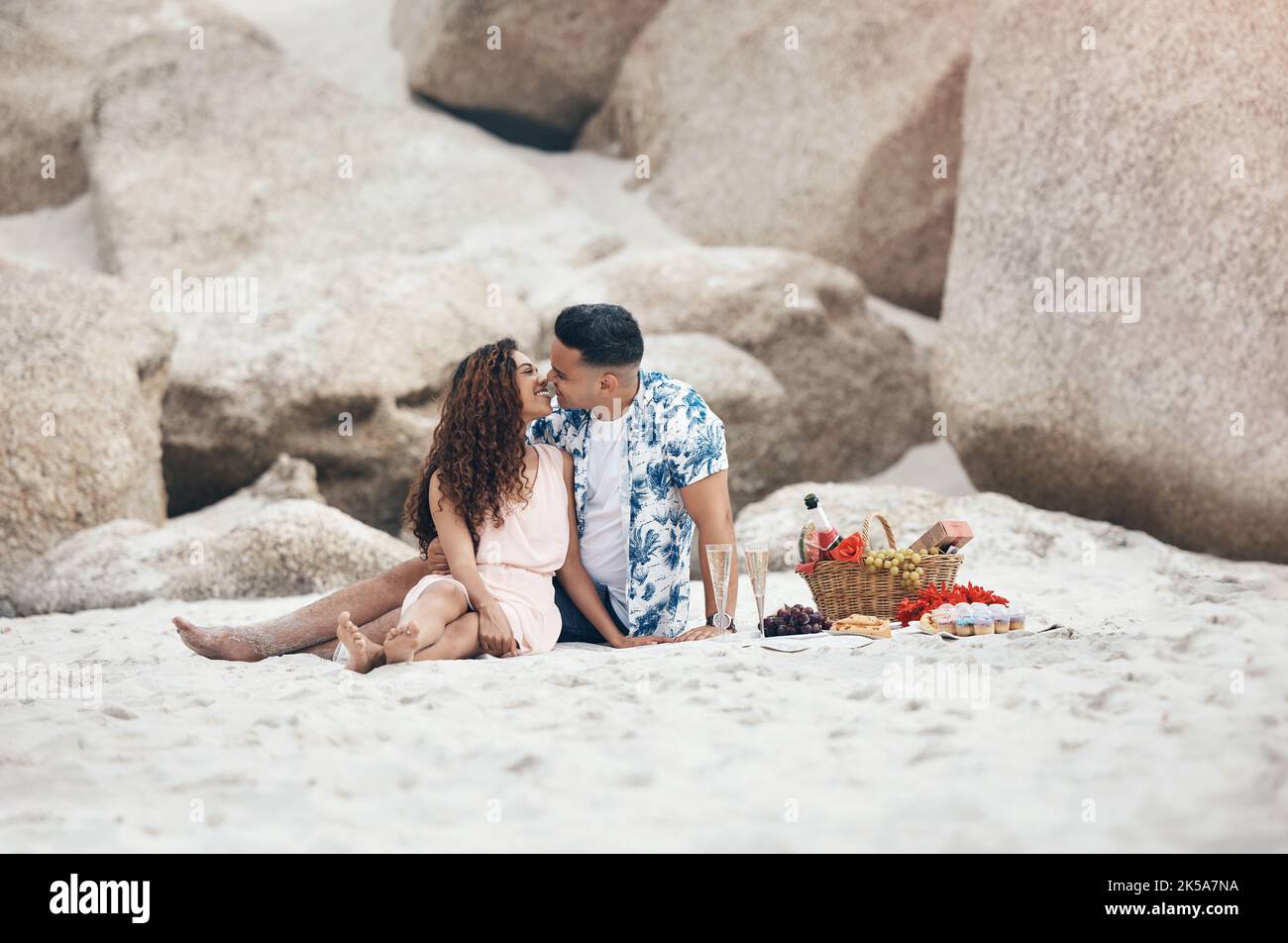 Coppia, spiaggia e amore al pic-nic, bacio e felice insieme sulla sabbia dalla roccia. Uomo, donna e sorriso con felicità sul viso con labbra, cesto e fiori Foto Stock