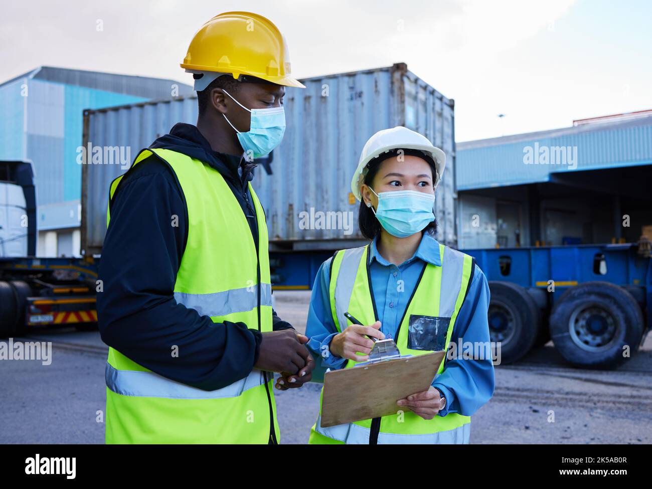 Covid, logistica e checklist dipendenti e maschera presso il cantiere, appunti e discutere il trasporto di merci. I dipendenti, uomini neri e donne asiatiche parlano Foto Stock