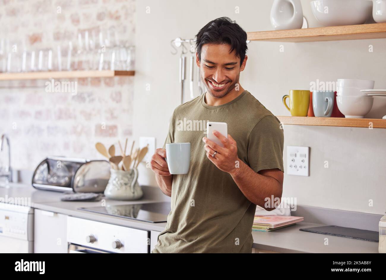 Telefono, caffè e social media con un uomo nella cucina di casa sua al mattino con un sorriso. Mobile, comunicazione e internet con un giovane maschio Foto Stock